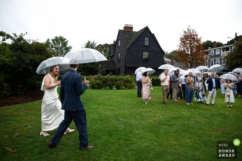 In the rain at The House of the Seven Gables in Salem, MA, a couple walks down an outdoor aisle, sharing umbrellas as they head to their wedding ceremony.