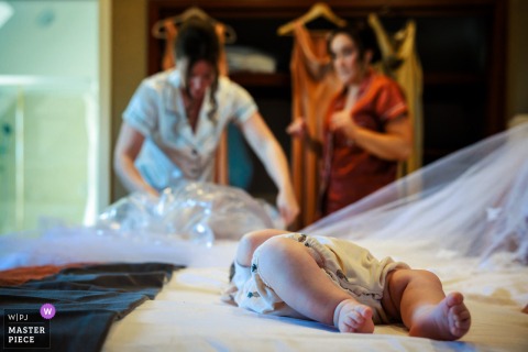 At a reception venue in Stirlingshire, Scotland, a bridesmaid's baby lies on the bed while the bride adjusts her veil in the background, capturing a quiet, intimate scene of preparation and new beginnings.