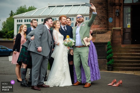 At Sherbrooke Castle Hotel in Glasgow, the bride and groom join their bridal party for a lively selfie in front of discarded shoes, capturing camaraderie and post-ceremony fun on their wedding day.