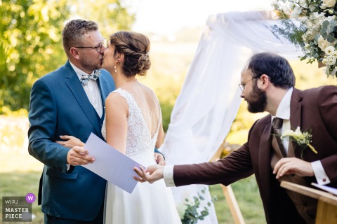 At Ferme Sainte Anne in Langres, the groom secretly hands his brother a file behind the bride’s back during their first kiss, capturing a playful and mischievous moment amid the romance of the ceremony.