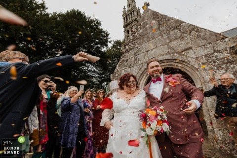Outside the Church of Ploeven in France, the newlyweds step into a shower of dried flower petals, their faces filled with joy as they exit the religious ceremony.