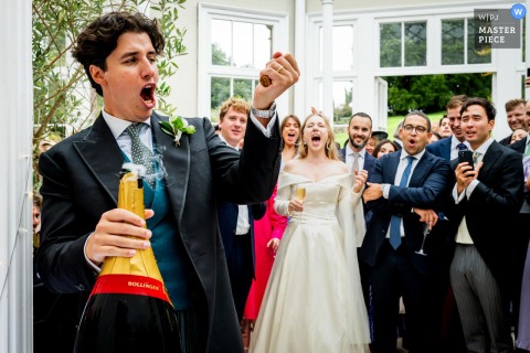 At Came House in Dorset, UK, the groom opens an impressive 15-litre bottle of Bollinger in front of his wife and guests, drawing a satisfying pop and a huge cheer. Captured from the kitchen, the image highlights celebration and excitement.