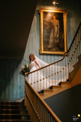 At St Giles House in Dorset, UK, the bride gracefully descends the staircase to greet her father waiting below, beneath the watchful gaze of an old master painting. Soft light from the overhead window enhances the rich stairwell colors.