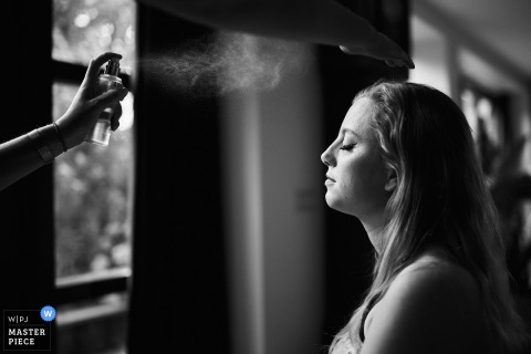 At the bride’s home, a black-and-white profile shot captures her getting makeup applied as a fine mist sprays across her face, highlighting the quiet anticipation and beauty of the wedding morning.