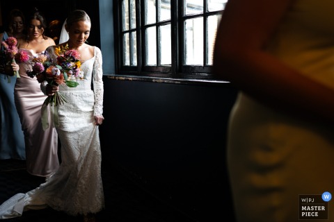 Dramatic Pre Ceremony Walk With Bride At The Bedford Balham At The Bedford in Balham, the bride and her party make their way to the ceremony, walking through a dim hallway illuminated by natural light from a window, creating a dramatic and atmospheric pre-ceremony moment.
