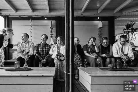 At a reception venue in Noord Holland, a candid scene reveals a contrast between two groups of guests—one lively and happy, the other looking bored—perfectly capturing the spectrum of emotions during the wedding celebration.