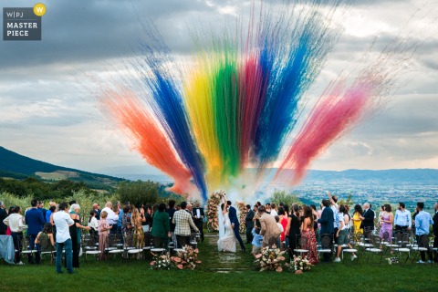 In Assisi, a vibrant rainbow smoke color explosion creates a dramatic backdrop behind an outdoor ceremony, filling the scene with joyful energy and a sense of celebration during the wedding.