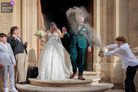 In Hérault, Occitanie, the bride and groom make their exit from the church, joyfully showered in lavender. A boy on the right enthusiastically tosses the flowers, getting the groom right in the face and adding playful energy to the celebration.