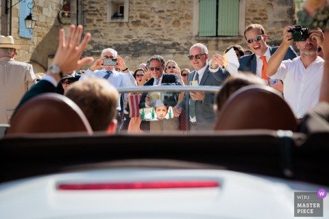 In Hérault, Occitanie, the bride and groom depart the church in a convertible to the applause of guests. A young boy watches them from the rearview mirror, adding a touch of curiosity and wonder to the joyful moment.