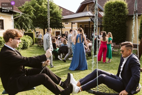 At Firlingerhof in Scharten, Austria, young men have fun on the kids' swings during the reception, their feet meeting mid-air under the trees on the grass. The scene captures carefree joy and playful celebration.