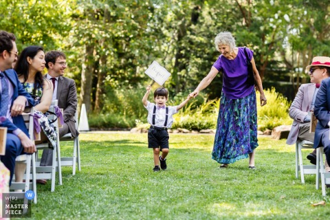 At an outdoor ceremony location in San Francisco, California, a young ring bearer walks down the grassy aisle carrying a pillow. The moment captures childhood charm and a sweet tradition on the wedding day.