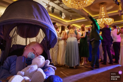 At Harvey’s Point Hotel in Co Donegal, a baby sleeps peacefully in a stroller amidst the lively party scene, while their parents dance nearby, capturing the gentle contrast between celebration and quiet family times.