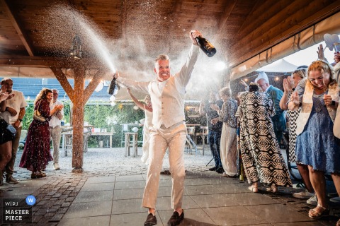 At a party location in Noord Brabant, the first dance comes alive as the groom joyfully sprays guests with champagne bottles, creating an exuberant and unforgettable atmosphere for the wedding celebration.