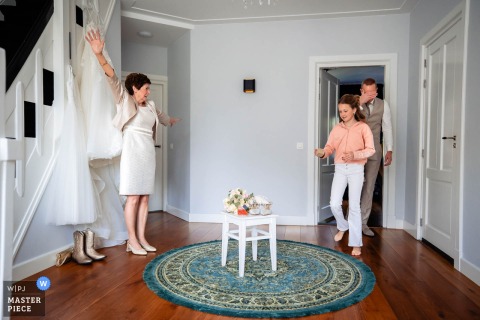 At home in Noord Brabant during wedding preparations, a woman uses her body to shield the hanging dress from view as a girl and a man enter the room, capturing a playful and protective getting-ready moment.