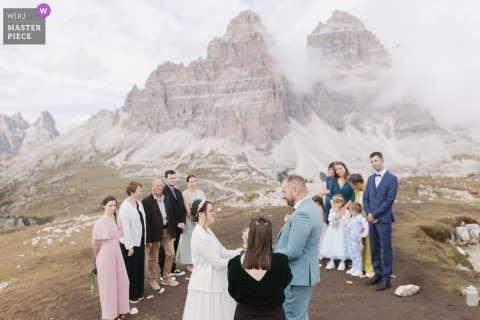 In the Dolomites, Italy, a wedding ceremony unfolds atop a mountain, with the newlyweds framed by dramatic, rugged peaks in the background. The breathtaking view adds wonder and grandeur to this unforgettable moment.