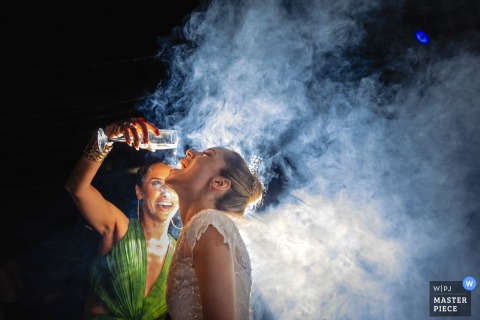 At the Cirağan Sarayı Kempinski Hotel in Istanbul, the bride and her friend share a lively moment as the friend pours a drink from her glass into the bride’s open mouth, surrounded by darkness and swirling fog. The scene exudes energy.