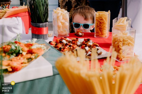 In Paris, a mischievous spirit shines as a boy wearing sunglasses peers at the hors d’oeuvres table, with only his head visible above the spread. The image captures youthful curiosity and playful charm.