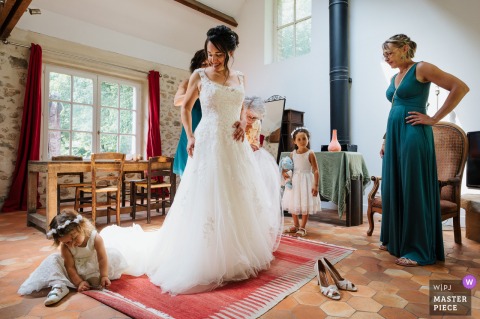At Moulin de Launoy, the bride receives help fastening her dress as a young girl plays with the sweeping train on the ground. The scene captures joyful preparation and innocence before the wedding ceremony.