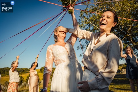 At Domaine de Baulieu in France, during the bouquet ribbon game, a witness playfully covers the bride’s eyes with her hand. Captured outdoors in the sunlight, the scene is full of anticipation and cheerful celebration.