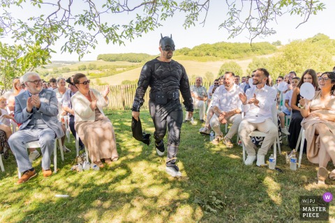 At Domaine de Baulieu in Gers, France, one of the wedding witnesses makes a memorable entrance dressed as Batman. Outside, under the trees and on sun-dappled grass, the lighthearted moment brings laughter to the ceremony.