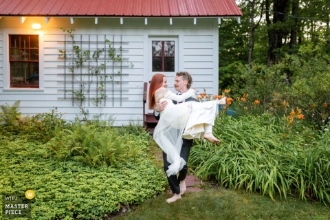 At a private residence in Newport, VT, the groom spontaneously sweeps the bride off her feet and carries her along a garden path near a building.