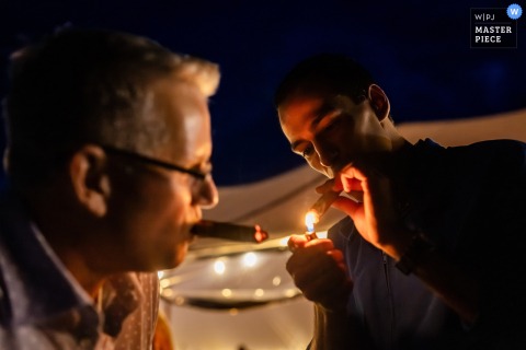 At a private residence in New London, NH, two wedding guests gather outside the tent at night to light up cigars during the reception. The scene captures camaraderie and celebration under the evening sky.