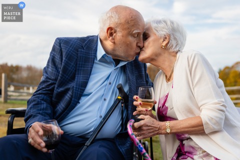 A sweet moment at a private residence in Hartland, VT, as an elder couple shares a kiss while sitting side by side on a bench outdoors during the wedding reception.