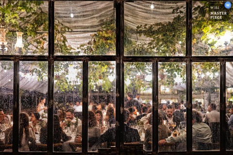 An atmospheric scene at a Cagliari, Sardinia reception, as unexpected September rain begins during dinner, creating a cozy ambiance. Captured through the glass windows, the scene beautifully contrasts warmth inside with the chilly weather outdoors.