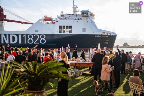 A festive outdoor scene at a Cagliari, Sardinia reception, as guests enjoy the dessert buffet under sunlight. A passing cargo ship joins the celebration, giving a friendly wave to the party, adding a joyful touch.