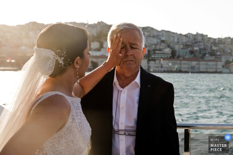 A tender moment in Istanbul, Turkey, as the bride gently comforts her father by the water, wiping away his tears in the sunshine—capturing heartfelt emotion and the warmth of their bond.