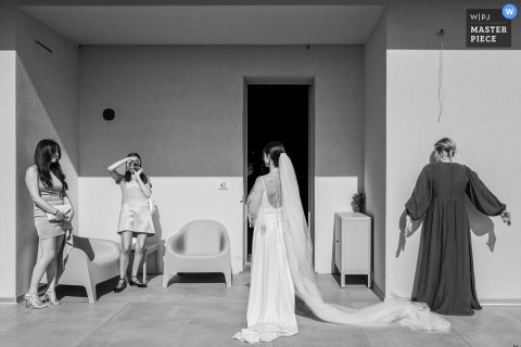 A playful behind-the-scenes moment in Cagliari, Sardinia, as the bride, ready for her day, asks a friend to snap a photo with a film camera—while another friend is caught trying to hide behind the wall.