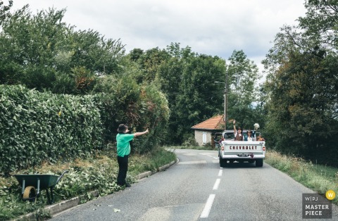 A charming moment at Chateau d'Arcine as a neighbor, gardening nearby, waves and cheers for the bride and groom as they head to their ceremony, passing by in an open car—capturing spontaneous community warmth.