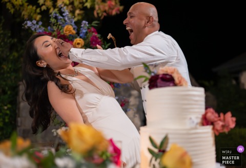 A lively and candid moment at a Santa Barbara Central Coast reception, as the bride leans back, surprised, getting a mouthful of cake from the groom, with a cake tower prominently in the foreground.