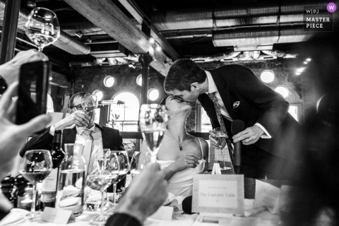 A romantic scene at Smiths of Smithfield, London, as the bride and groom share a kiss at the end of the speeches. Captured in black and white, the image is artistically framed through soft-focus, raised glasses.