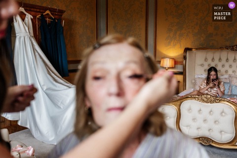 A candid moment captured at Rise Hall, Yorkshire, England, as guests prepare in the morning. A woman in the foreground gets her eye makeup applied in soft focus, while the bride’s dress hangs in the background—beautifully documented.