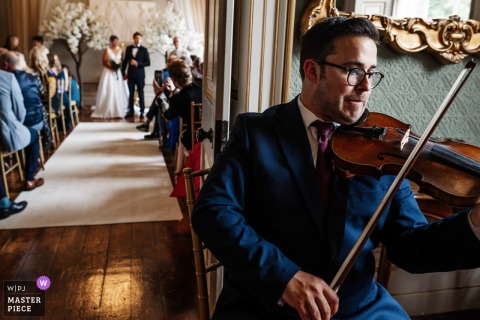 A memorable moment captured at Rise Hall, Yorkshire, England, as a violinist, in sharp focus at the front right, plays while the bride and groom prepare to leave the ceremony—beautifully documented.