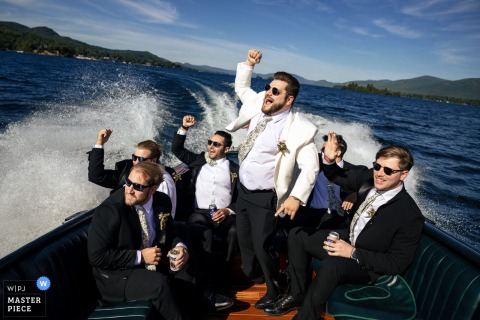 A lively moment captured at The Erlowest on Lake George, NY, as the groom and his wedding party celebrate on a fast-moving boat, cheering with fists overhead and leaving a wake behind—a raucous scene beautifully documented.