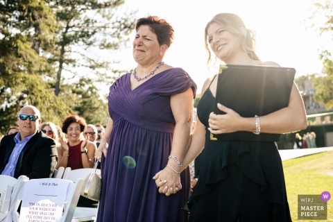 An emotional moment captured at The Erlowest on Lake George, NY, as the groom’s mother, visibly moved, walks down the aisle with his sister, who also officiated the outdoor ceremony under trees and sunlight.