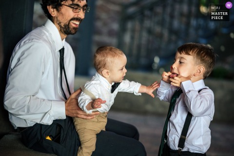 A joyful moment captured at The Erlowest on Lake George, NY, as a man holds a toddler on his lap and they share a playful interaction with a small boy during cocktail hour, beautifully documented.