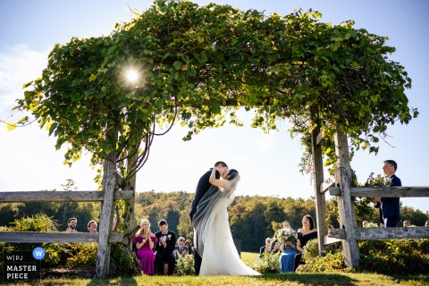 A memorable moment captured at Mountain Top Inn in Chittenden, VT, as the bride and groom share their first kiss beneath a foliage-covered arch, with the sunburst and clear sky over their ceremony.