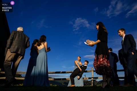 A lively moment captured at Mountain Top Inn, Chittenden, VT, as wedding guests snap photos during cocktail hour. A playful woman dips a man under afternoon light and a deep blue sky, beautifully documented from a low angle.