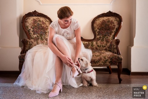 A charming moment captured at Villa Marchese De Fabris, Gorizia, Italy, as the bride sits on a vintage couch putting on her shoes, while her little dog seeks cuddles—symmetrical framing beautifully documents this sweet scene.