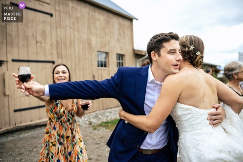 A candid moment captured at Isham Farm in Williston, VT, as the bride receives a hug and guests carefully keep a glass of wine, held by the hugging man, away from her dress—beautifully documented.