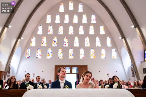 An emotional moment captured in Trieste, Italy, as the bride sits at the altar with her groom, overcome with emotion during the ceremony, beautifully documented with symmetrical framing and a massive stained glass arch as the backdrop.