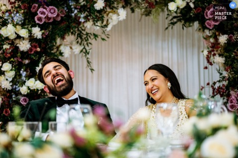 A joyful moment captured at Offley Place, Hertfordshire, UK, as the very happy couple sits at the head table, laughing during a speech, beautifully framed under an arch of florals.