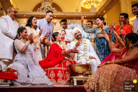 A festive moment captured at a wedding hall in Wolverhampton, UK, as a large group enjoys fun, traditional Hindu wedding games. The low camera angle documents the lively atmosphere and cultural celebration.