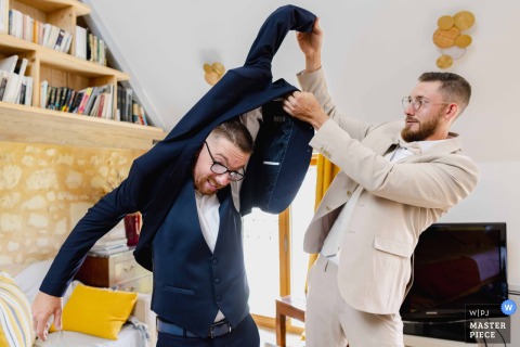 Trouble With A Jacket In Sainte Maure De Touraine, France, Leads The Groom To Gratefully Accept His Best Man’s Help. In Sainte Maure de Touraine, France, a groom has trouble putting on his dress jacket, gratefully accepting help from his best man before heading out to greet guests.