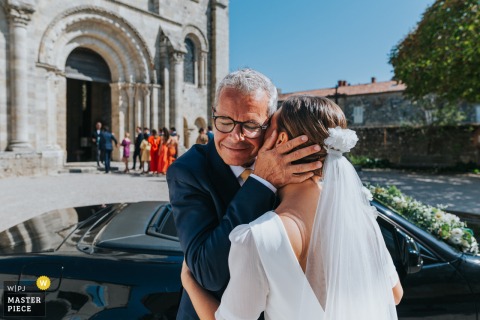   At Église Notre-Dame de Moirax, a touching moment unfolds as a father gives his daughter a loving kiss just before the ceremony begins, symbolizing support and affection on her wedding day.