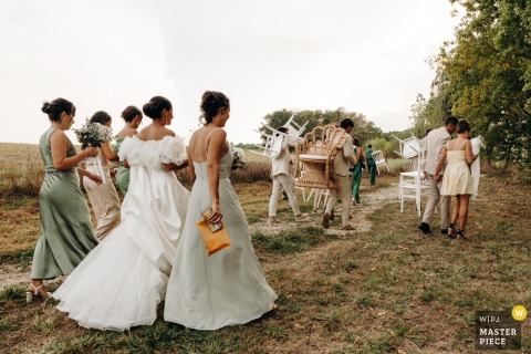 Guests Share Community Spirit Carrying Chairs Together After A Ceremony At Chateau In Gers, France. After the ceremony at Chateau in Gers, guests pitch in, carrying chairs from the event space, sharing in the togetherness and community spirit of the celebration.