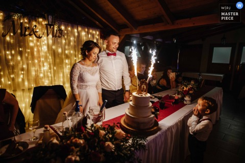A Sparkling, Joyful Moment Emerges By The Wedding Cake In Budapest As The Couple Stands Surrounded By Lights And Sparklers. Surrounded by sparklers and glowing lights at the reception venue in Budapest, the couple shares an intimate, joyful moment by their wedding cake, capturing the festive spirit.